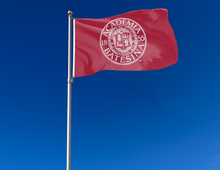 Load image into Gallery viewer, Red 3x5 foot wide Bates College Seal Flag with two metal grommets attached to flagpole as it waves in the wind
