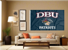 Load image into Gallery viewer, Blue 3x5 foot wide Dallas Baptist University Flag with DBU Patriots logo and two metal grommets covering the tan wall of a living room with couch and throw pillows
